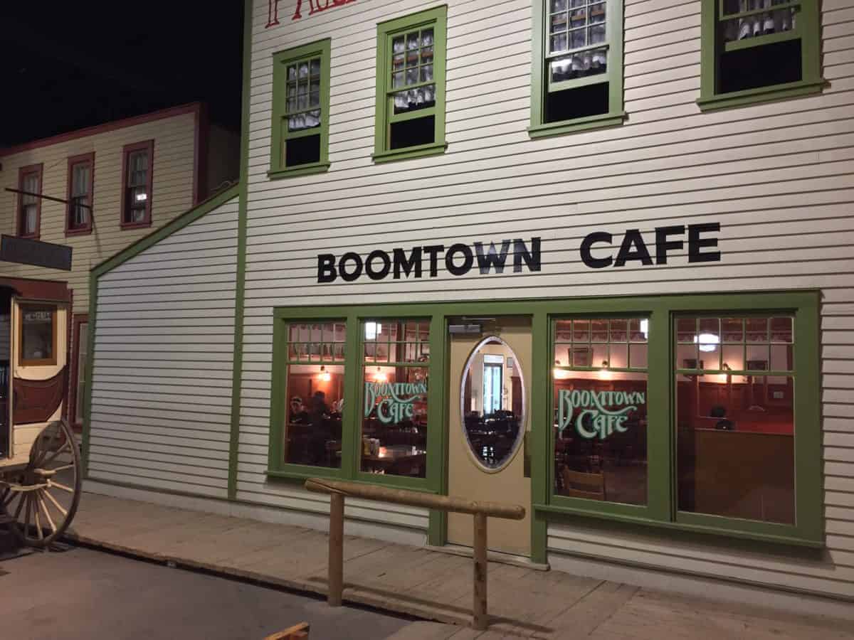 Front of Boomtown Cafe large white building, with green trimmed windows to peer into at street level and a hitching post out front.