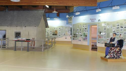 Carved figures of a woman and and a man on exhibit near a brown sod house.