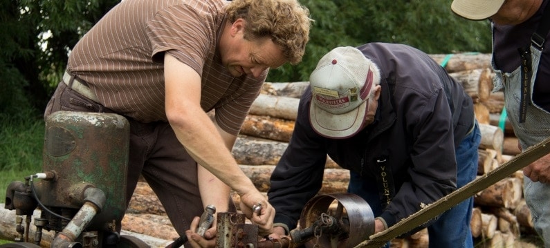 Two volunteers use tools to work on parts of an antique gas tractor