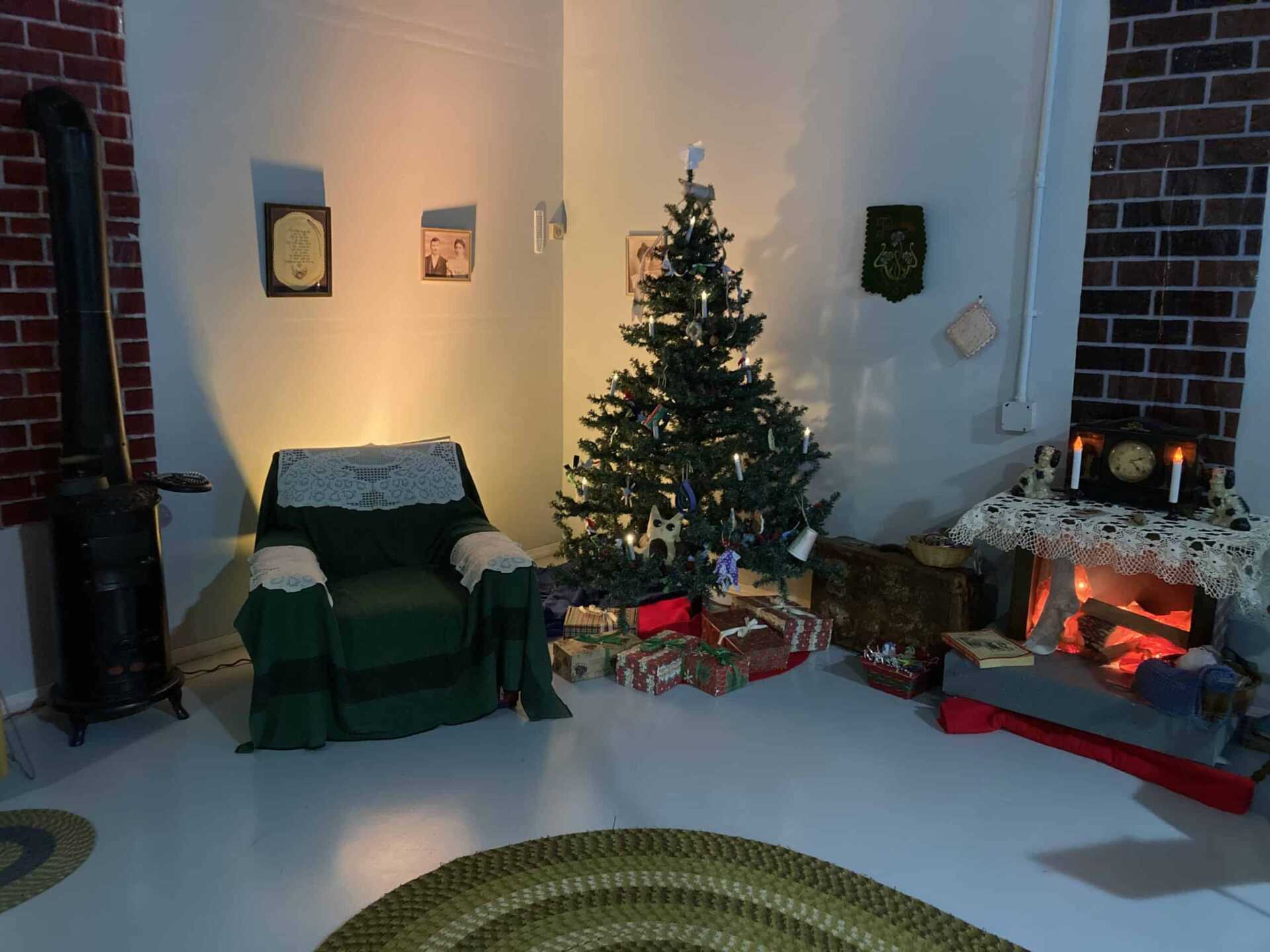 Cozy living room scene with a glowing (fake) fireplace, green braided rug ont he floor, and armchair beside a decorated Christmas tree.
