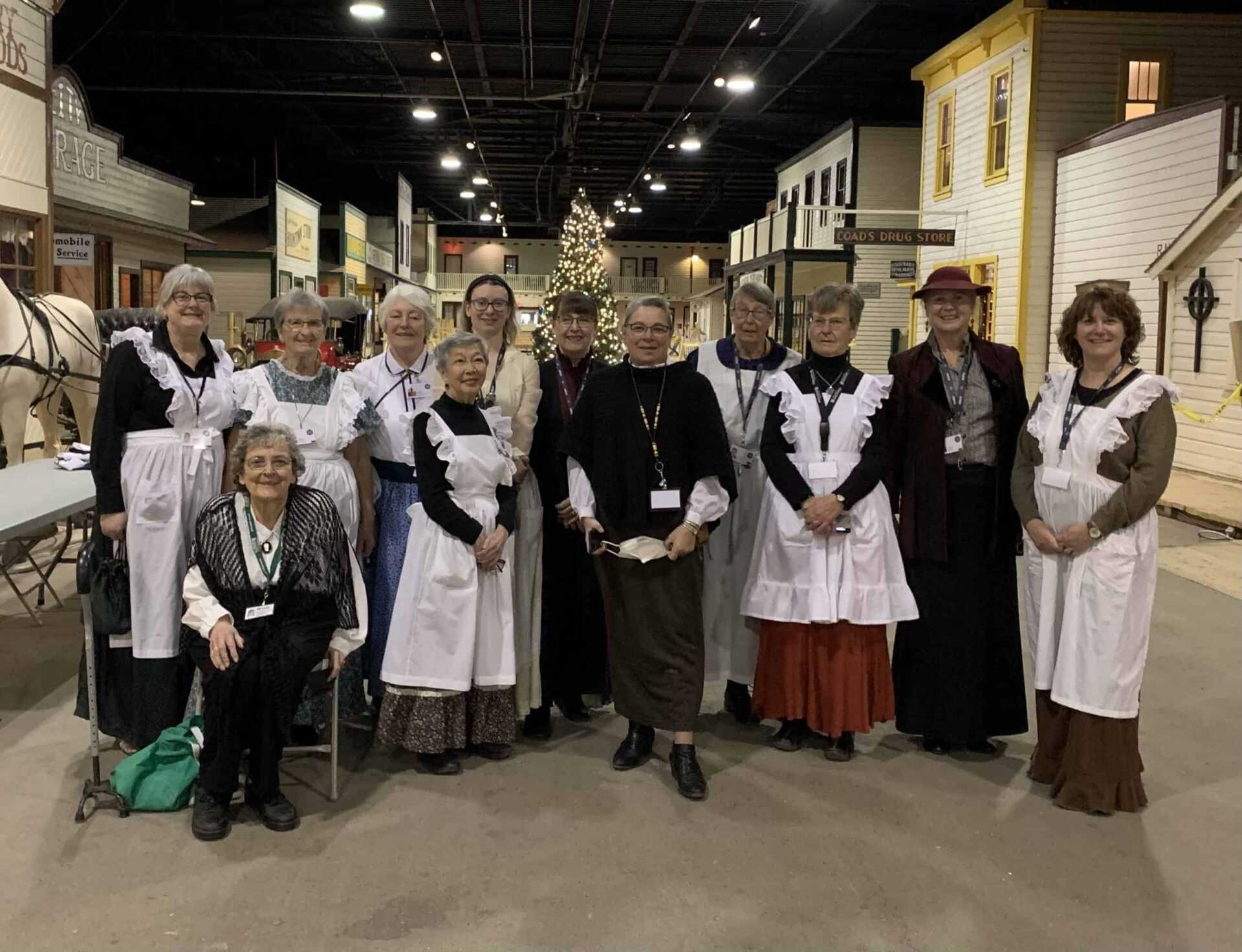 Group of women, most wearing aprons, posted as a group in front of a large Christms tree