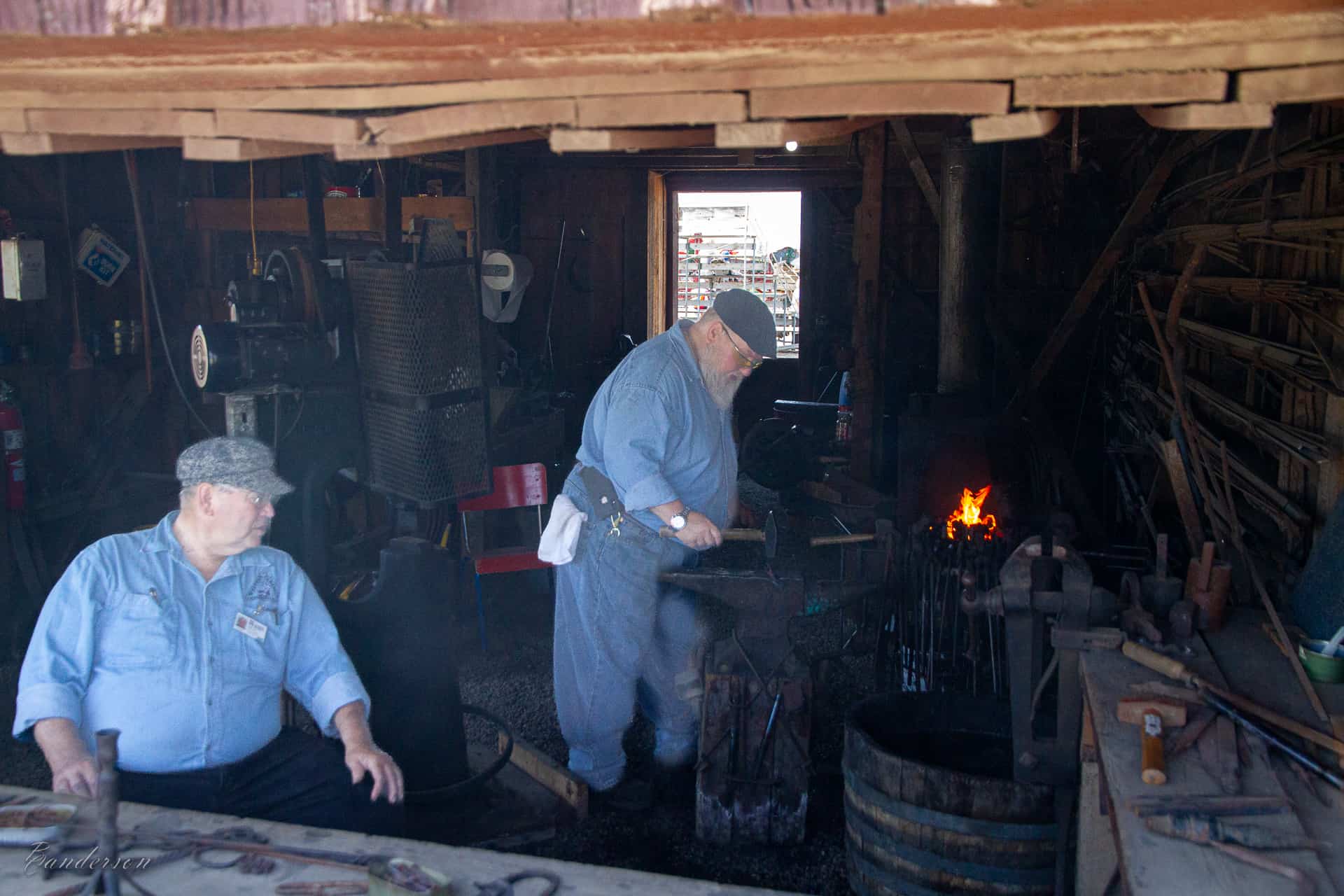 Two men inside a blacksmith shop. One sits near the front. The other leans over a lit forge.