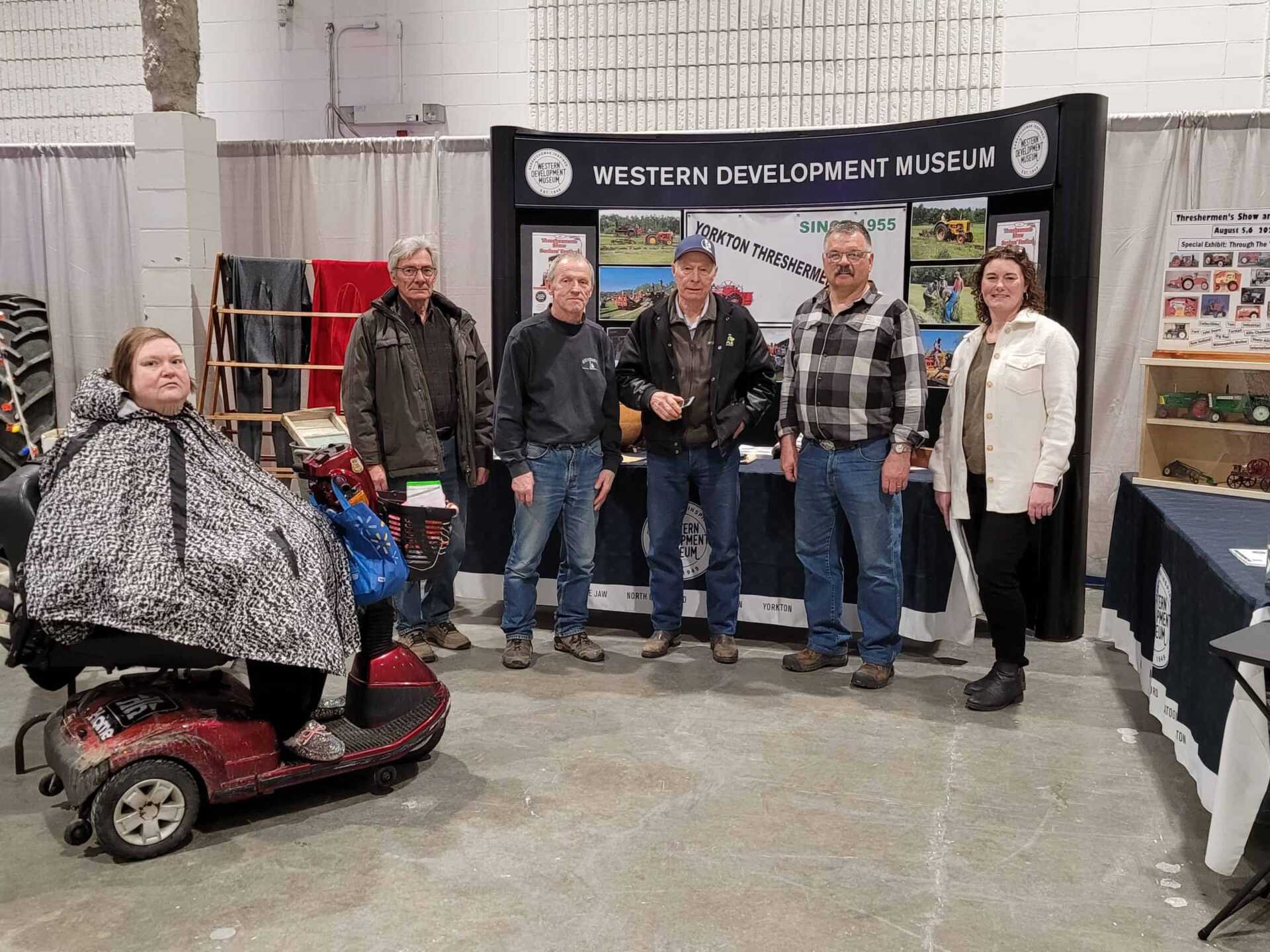 Group of people standing and one sitting in front of a banner reading "Western Development Museum"