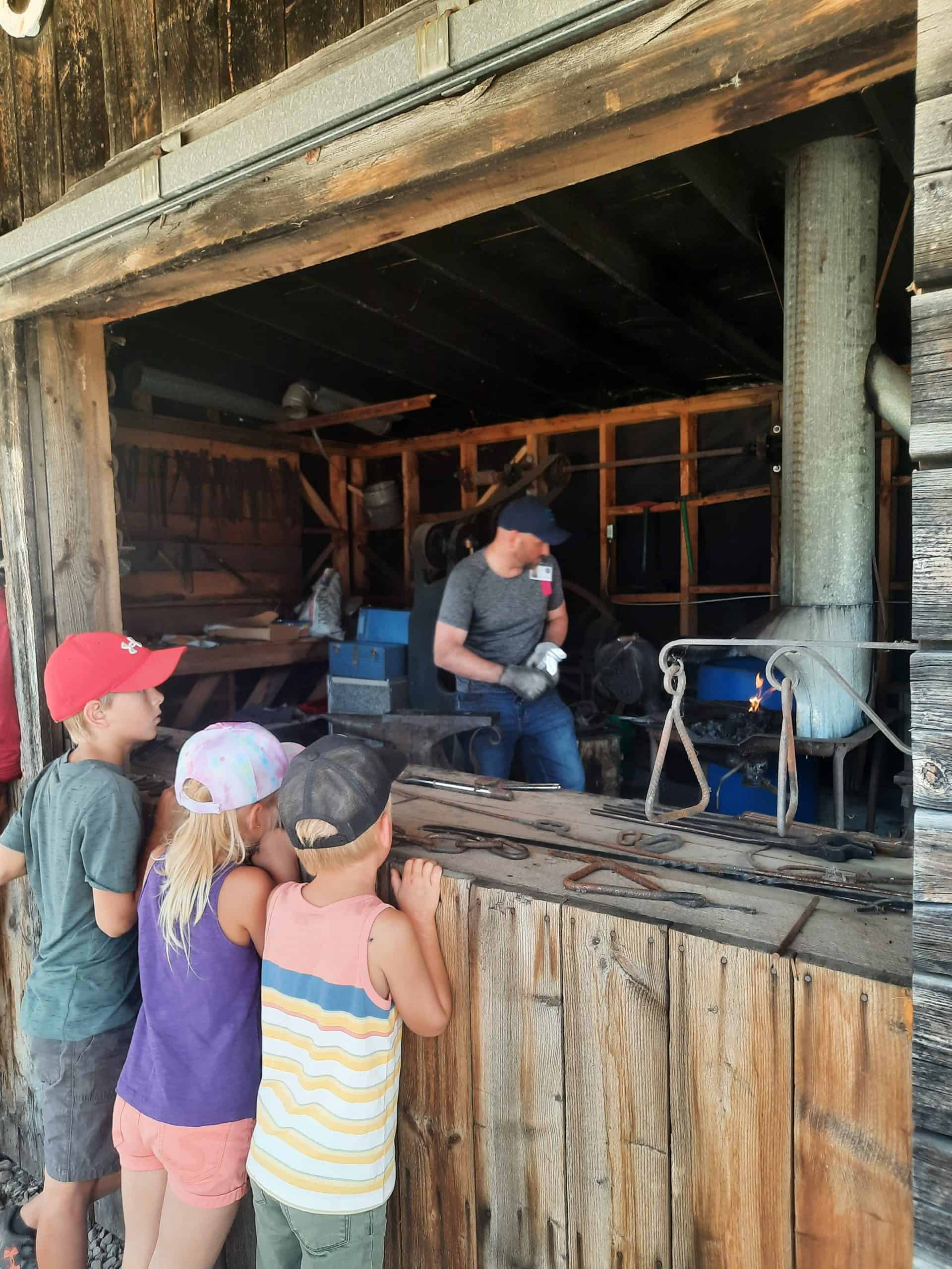 Three kids peer into a window to watch a blacksmith at work