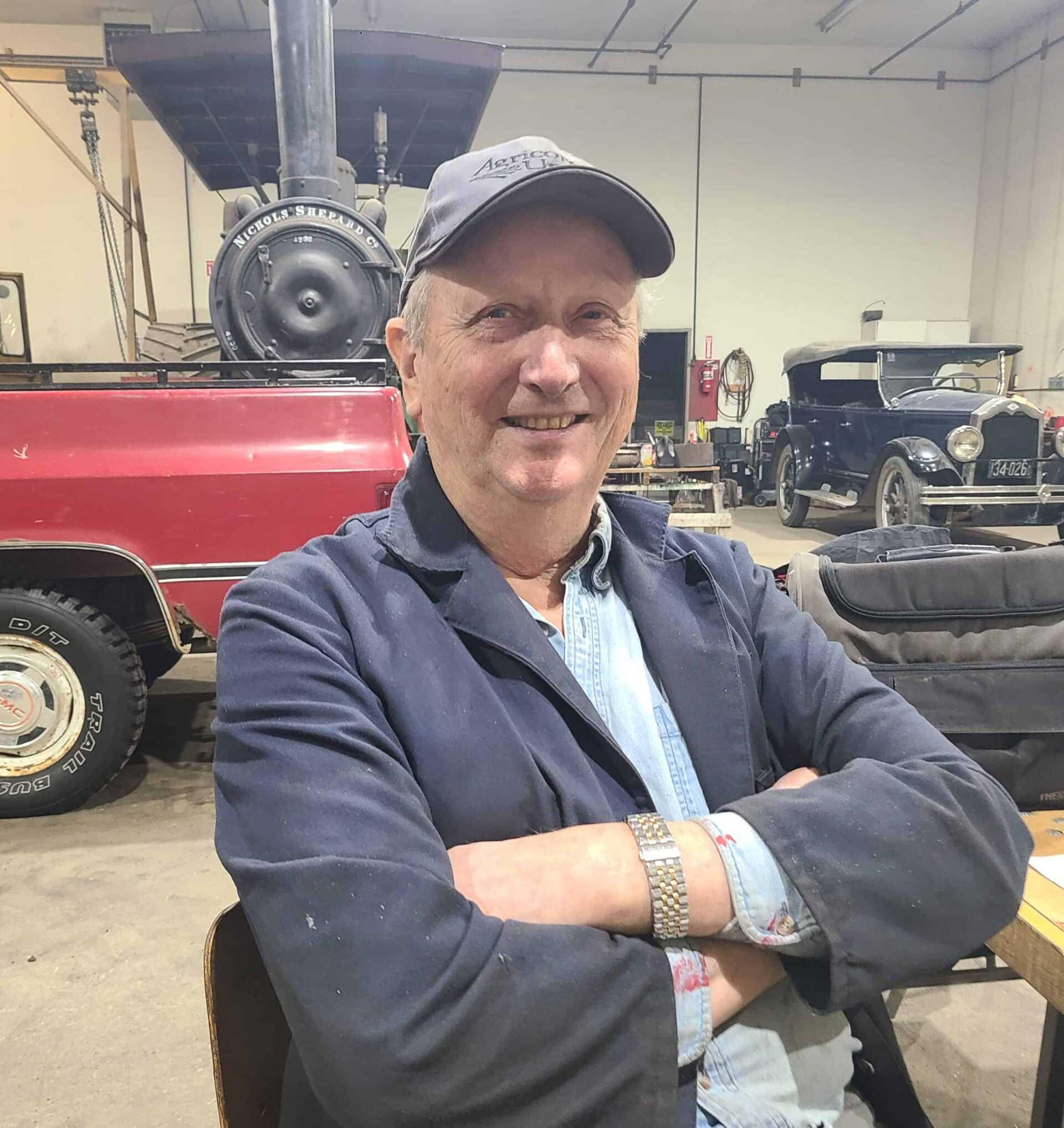 Man with big smile sits, arms crossed, in front of a steam traction engine and bright red truck