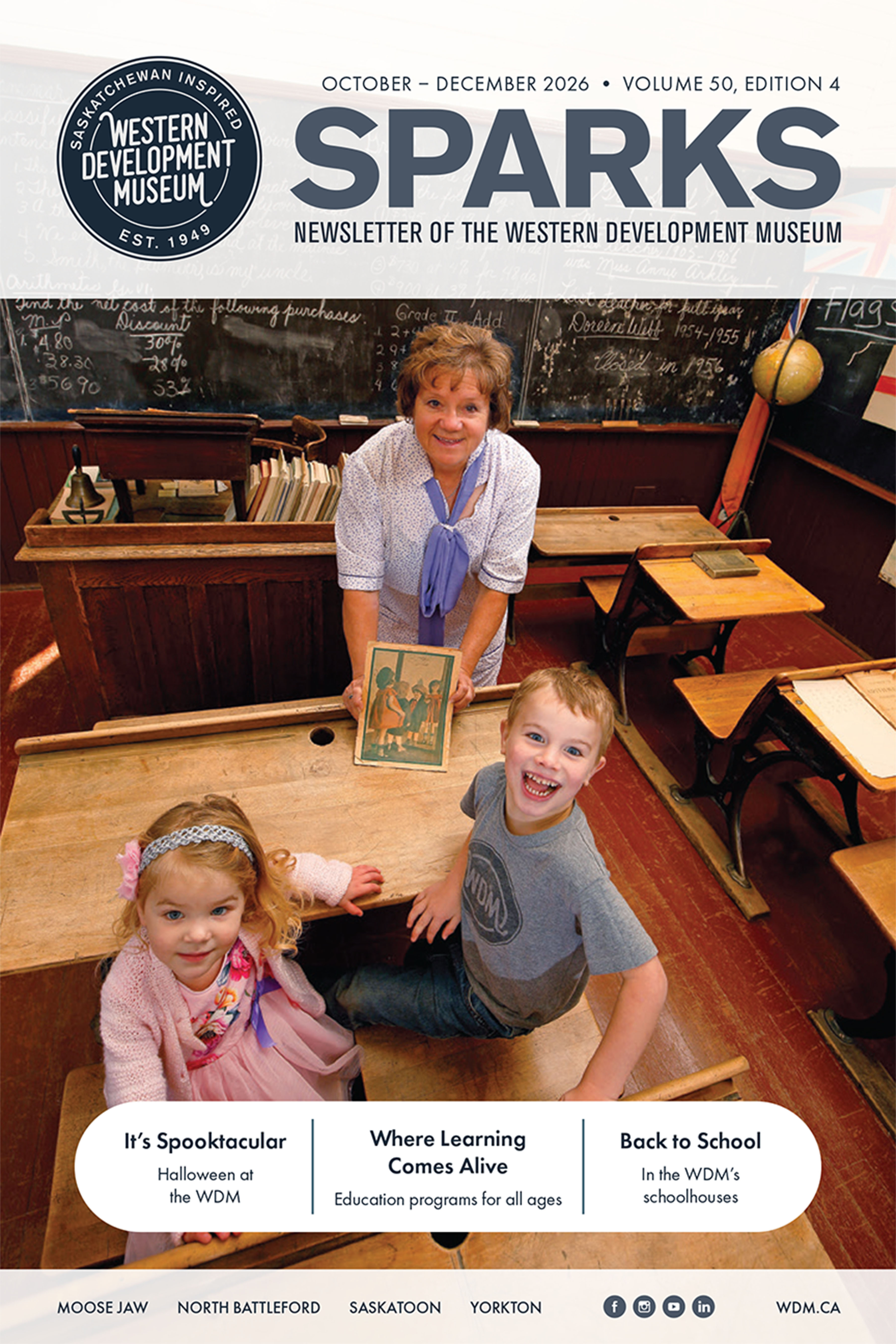 A woman shows a photo to a young girl and boy who are sitting in a wooden desk inside a one-room schoolhouse